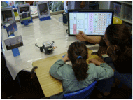 Figure shows a child facing a computer screen with a communication grid. The grid includes vocabulary cells as well as robot command cells (e.g. forward, backward, left, right). On the same table where the screen is, it is possible to see a small car-like Lego robot with a gripper and a pen attached, along with educational items to be manipulated by the robot. 