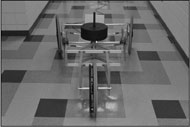 Picture of the misalignment cart. The cart has an aluminum frame with joints that allow camber and toe misalignment to be controlled. The cart has two twenty two inch wheels in the front, facing away from the camera, and one wheel in the rear at facing the camera. The wheels are in a significant toe out misalignment angle. In the mid-section of the cart are two large weights on a post.  