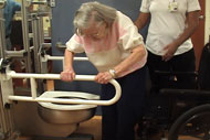 In a bathroom lab with adjustable fixtures, a female older adult grabs the bilateral toilet grab bars with two hands after ambulating from a wheelchair to transfer to the toilet. A caregiver assistant stands in the background ready to assist. 