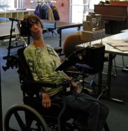 Client sits in her chair with the device mounted to the wheelchair crossbar behind her permanently mounted communication board.  The pan control button is visible beside her left knee.  The trigger control button is not visible, but is beside her left hand.  These controls could be positioned anywhere around her that would be most convenient.