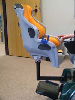 The picture shows the baby seat and mounting hardware attached to the client's wheelchair. The seat is facing the client. 