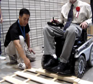 Pictures of two wheelchair users demonstrating driving with the “Patrafour” over  2inch obstacles-one obstacle is a step and the other obstacle  is a piece of wood