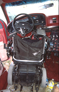 Rear view of driver’s wheelchair in secured in docking tiedown device after the frontal crash.  The docking device is deformed such that the wheelchair is tipped slightly toward the right side of the van.  The tri-pin steering device is visible on the steering wheel.