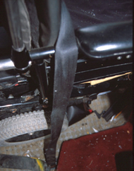 Close-up photo showing routing of the vehicle-anchored shoulder belt over the right armrest of the wheelchair to the anchor point on the floor.