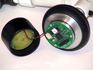The photograph shows a miniature data logger which is about the size of a baby food jar.  The plastic enclosure has been opened; the plastic cap contains the battery.  The printed circuit board is visible on the aluminum base.  The aluminum base also forms the hub of the three inch caster wheel.