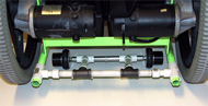 This photograph is taken from the rear of an electric powered wheelchair.  The 24 inch back wheels are partially visible.  A structure constructed of white plastic tubing is attached to the lower wheelchair frame just an inch or two above the floor. At the front of this tube structure is an aluminum axle about twelve inches wide; on each end of the axle is mounted a miniature data logger.  Each data logger caster is located just inboard of a drive wheel.  The data logger axis is aligned directly under the drive wheel axis.  This is done to prevent side scuffing of the data logger casters during turns.