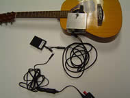 Image shows the overhead view of the entire device with connections. An AC/DC converter is just visible and is connected to a rectangular pedal through a 1/8” mono connection. The pedal is then connected to the strumming component through ¼” stereo connections. The strumming component is mounted to the guitar face. 