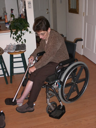 The image shows the final device in use by our client as a sock aid.  The string is threaded through the bracket on the back of the heel sleeve and the sock is stretched over the top of the shoehorn.  Our client is using her reacher to place the sock opening over her toes and will then use the string to pull the sock onto her foot.  