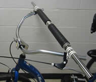 Photo 2 is a closer view of the custom handlebar and is meant to provide a better view of handlebar parts, including the hand grips, rotating cylinder, support shaft, shaft collars, and clamps. The metal bracket used to fasten the brake cable to the trike frame is also seen at the front of the trike’s existing handlebar. 