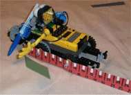This figure shows a Lego Robot configured like a car with wheels.  There is a 30 cm ruler attached to the wheels of the car.  The robot can be moved forward and backward to measure the length of the paper cut out of a parallelogram that is laying on the table next to the robot.  There is also a marking pen mounted to the front of the robot on a mechanism that can be moved up and down.   