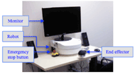 The figure shows the upper limb rehabilitation robot. It is based on top of a desk. It has an extruding two-link arm with a half-sphere end-effector. On top of the robot is a monitor to display the Graphical User Interface. On the right side of the robot is an emergency stop button. On either side of the robot are speakers.  