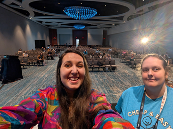 Two women taking a selfie in front of a conference room filled with people