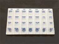 This photograph shows a pill box. The pill box has seven columns labeled SUN, MON, TUES, WED, THURS, FRI, SAT and four rows labeled MORN, NOON, EVE, BED, making 28 total compartments. The pill box is white with raised blue text and Braille markings.