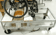 This photo illustrates how the AMPS is used in conjunction with the dynamometer.  The AMPS is loaded atop the dyno frame, with the casters anchored into a bracket while the drive wheels rotate with little or no slip over the large dynamometer drum.