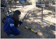 Photo of a woman at a campsite squatting down to measure the grade of the clear ground space in front of a cooking grill with a SmartToolTM 