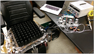 A photograph of the entire apparatus is shown. The modified cushion is on a power wheelchair, an air compressor is on the ground next to the power wheelchair, and a breadboarded electronic controller is on a tabletop next to the power wheelchair.