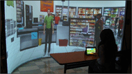 Figure 2: A picture showing a user who is training on money management skills in a virtual grocery store. The user trains on recognizing coin and bill amounts subtask by interacting with a touchscreen computer. The touchscreen computer represents the cash register and the virtual world is projected onto the 180⁰ curtain display. 