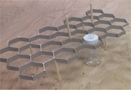 The Honeycomb Helper is a device to help workers with disabilities package pharmaceutical bottles in a cardboard box to achieve an alternating, client-specified pattern. The device includes 32 hexagonal holes that help guide bottle emplacement, bumpers to guide the positioning of the remaining 18 bottles, five supporting legs, and two handles.