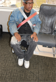 Figure 1 Right is a front view of a non-ambulatory man seated in a Columbia Aislemaster boarding chair with shoulder straps. One strap is red the other strap is white. The straps create an “X” pattern across the man’s chest.
