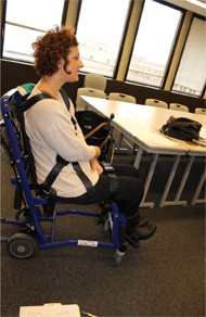 Figure 2 Left is a side view of a non-ambulatory female seated in a Staxi boarding chair. Positioning straps can be seen going from the abdomen to the rear of the seat and to the front of the seat creating a triangle. A shoulder strap can also be seen. All of the straps are black.
