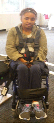 Figure 2 Right is a front view of a non-ambulatory female seated in a Staxi boarding chair. Positioning straps can be seen going across the chest of the person forming an “H” pattern. The top of the “H” is right below the shoulders. All of the straps are black.