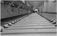 The picture shows multiple modular rough-surface units placed in series on a floor in a hallway of a building, making a 30-meter straight rough pathway. A modular unit consists of wooden boards placed in parallel with space in-between. Both ends of each board are attached to two parallel PVC pipes using rubber strips wrapped around the pipes and stapled on the board.  