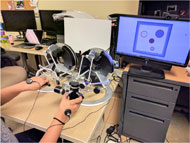In the image, there is a blindfolded user exploring an image with two haptic devices. Each of his hand is holding one haptic device. There is also a vibration tactor attached on the back of each hand.