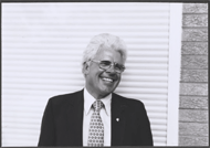 A black and white photograph of Fred Sammons, a man with white hair and glasses, wearing a dark suit and tie, in front of white siding.  He’s chuckling to himself.