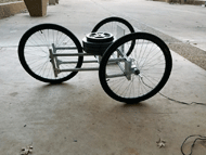 Picture of the rolling resistance cart showing an aluminum frame approximately 3 feet wide and 3 feet long.  The frame holds three wheel chair wheels like a reversed tricycle with two wheels in the front and one in the back.  The cart has a load cell attached to the front to pull the cart forward and a metal plate mounted as a target for the laser sensor.  Barbell weights are pile in the center to exert weight equally to each wheel. The cart is placed on top of smooth, level concrete.  