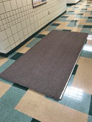 Picture showing a 4-foot by 8-foot melamine board with commercial carpet adhered to the top surface.  The picture also shows the shiny smooth vinyl tile floor of the hallway where rolling resistance is tested as a “smooth floor” surface. 