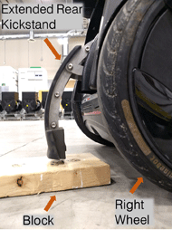 Photo of kickstand block testing setup. Close up of extended rear kickstand mechanism. Right wheel is in foreground. A wooden block is placed on the floor where the kickstand is about to land. 