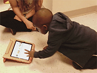 A young child with expressive communication challenges uses the yCAT website on an iPad. He kneels on the floor with a researcher nearby and touches the screen while looking at the tablet with engagement.

