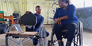 : Figure 2 is a picture of a technician showing a wheelchair user how to remove and fold the new seating unit while located at the clinic site. The technician is kneeling in the photo to be at eye level with the wheelchair user when discussing the new seating units components. 	