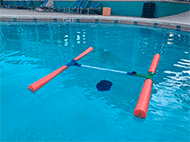 In this picture, the device is unfolded and deployed in the center of a swimming pool. The device's four orange buoys are perpendicular to the central bar, giving the device an H shape as it floats on the surface of the pool's water. Submerged beneath the water, nine black weighted plates (apparent weight of 18lbs) hang from a string tied around the middle of the central PVC axis of the device. Weights were added to test buoyancy capabilities of the device. Results showed that the joint regions and middle portion of the device were submerged below the water. The polyethylene foam-wrapped appendages of the device floated on the surface of the water. The middle segments of the device were not wrapped in foam; thus, the central axis of the device is slightly submerged underwater. 