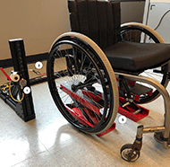 Laser measurement system for toe and slop. (1) Laser, (2) Constant Force Springs (3) Lift Mechanism.  A picture of a manual wheelchair lifted slightly off the ground on two small jacks (comprising the lift mechanism), and constant force springs hooked onto the rear wheels to induce toe.  The lasers are mounted on to a metal frame 33 inches wide, between which the wheelchair is placed.  The lasers accurately measure the distance to the wheel on both sides.