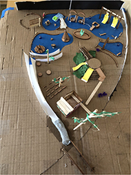 Photo of 3D map, which is a scale model of the Magical Bridge Playground. The model is constructed out of cardboard, paper and clay and includes slides, swings, a long curved ramp and other playground features.