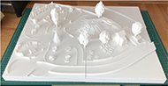 3D-printed model of Magical Bridge Playground, printed in white plastic. The model is very realistic and shows a variety of playground features such as trees, swings, slides, walls and a small building.
