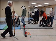 Figure 1. The students and spotters performing the soft-surface skill while simulating wheelchair users moving forward on gym mats and simulating caregivers pulling the wheelchairs backwards over gravel. The instructor is holding a rake used to deal with displaced gravel.