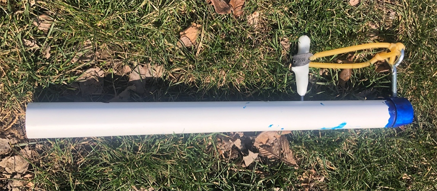 white PVC pipe with a blue end. Pipe is laying on the grass with an eye hook and handle attached via slingshot band.
