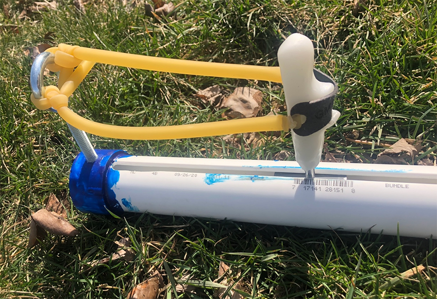  Yellow slingshot band tied around eye hook bolt at the blue launching end of device. Slingshot band is linked to the white instamorph handle in the white PVC pipe.