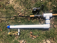 fishing rod attached to white PVC Pipe with a tee joint and 90 degree elbow with a white handle and eyelet hook connected by slingshot band at the end.