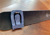 The image displays a close-up view of the left-side of the SlapBra device. The SlapBra is lying flat on the table, with the yoga mat slap-band straightened. The left half of the screen displays the blue tab attached to the yoga mat via a velcro tab; The tab is composed/made of thermoplastic material and appears with an upside down “U” cut-out to create a hook for the bra fasteners to attach onto.