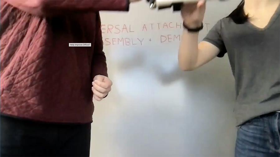 How to Use the Universal Attachment Part 2. This picture shows 2 people using the Universal Attachment. (Part 2) The person assisting the client lifts the device towards the users mouth in the case of feeding.