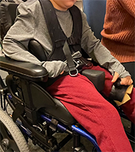 : figure of a deidentified patient in a wheelchair using a prototype Orthoplast hand anchor. The device is placed around his left thigh and he is holding it with a gross grasp to stabilize his movement. 