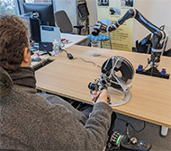 A photograph of a tetraplegic user operating the haptic controller as a 3D joystick to position a robotic arm in order to pour a typical drinking cup.