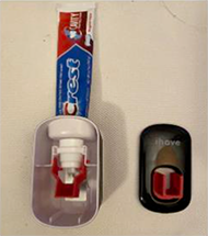 The toothbrush buddy dispenser is shown with the front cover taken off. Inside the dispenser the red lever is attached into the dispenser and the white suction piece is shown. A red and blue toothpaste tube sticks out of the top of the dispenser. To the right, the black lid with the red track attached is shown.