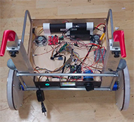 : A view of Shelbytron with the dog and electronics box top removed. Most of the space inside the electronics box is taken up with a large ribbon cable and the wires that branch off from it. The battery and speakers are towards the front, the Teensy is towards the center, and the power switch and motor controller board are at the back.