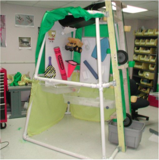 Figure 1 shows the front view of the Pouring Palace. On one end, the lifting and cranking mechanism is mounted to the outside of the PVC frame. On the adjacent side, a magnetic whiteboard is mounted to the interior of the frame. Various gutter parts and tubes are magnetically stuck to the board. On the side opposite the lifting mechanism, a white pegboard is mounted to the interior of the frame. Various toys are hung from hooks inserted into the pegboard. A green and yellow fabric tent veils the entire structure.