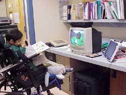 This photograph shows a student using his DynaVox 3100 (on right side of counter top) to play PlayStation2 (black box on top of TV). The student is using a spec switch, which is secured in his right hand with a velcro strap, to perform auditory single switch scanning. 