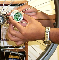 Figure 1 is a photograph of a hand holding the new miniature data logger and inserting it between the spokes of a manual wheelchair wheel. The miniature data logger is round about the diameter of a pocket watch and approximately one and one half inches thick. 